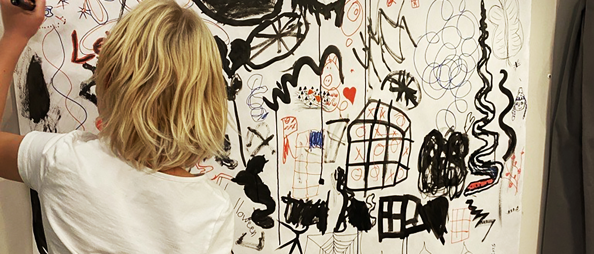 Ett stort papper på en vägg med målade figurer på. I förgrunden står ett barn och målar på väggen.